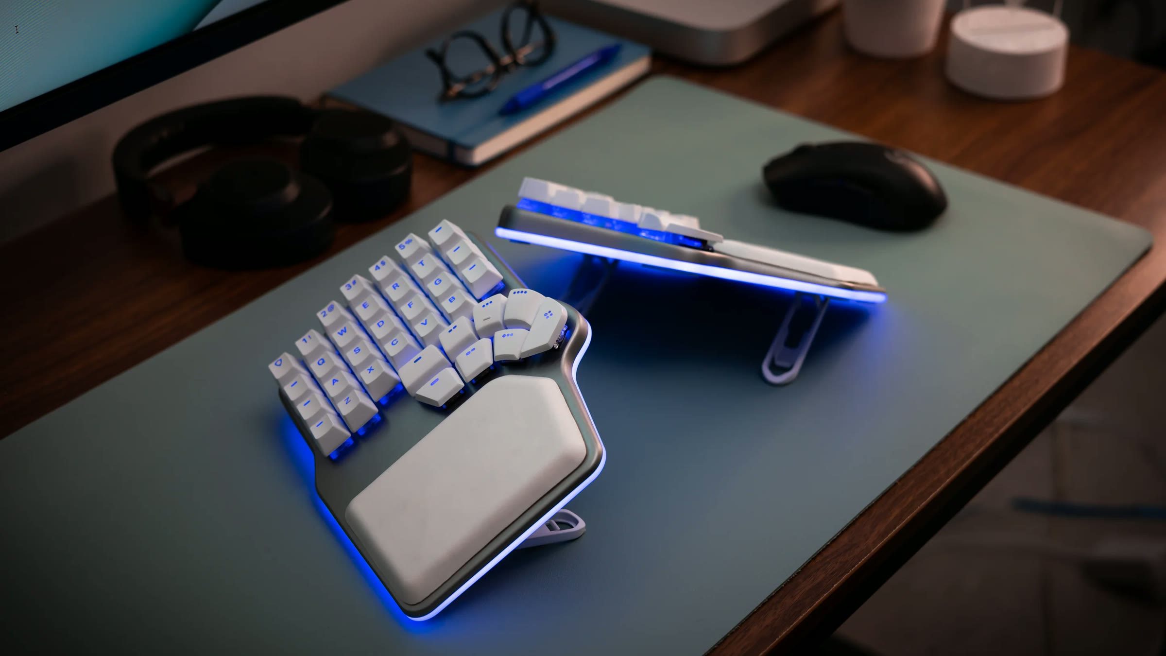 A Dygma Defy split keyboard on a blue-grey desk mat with blue underglow, one half flat and the other tilted on its stand, next to a mouse, headphones and a notebook.