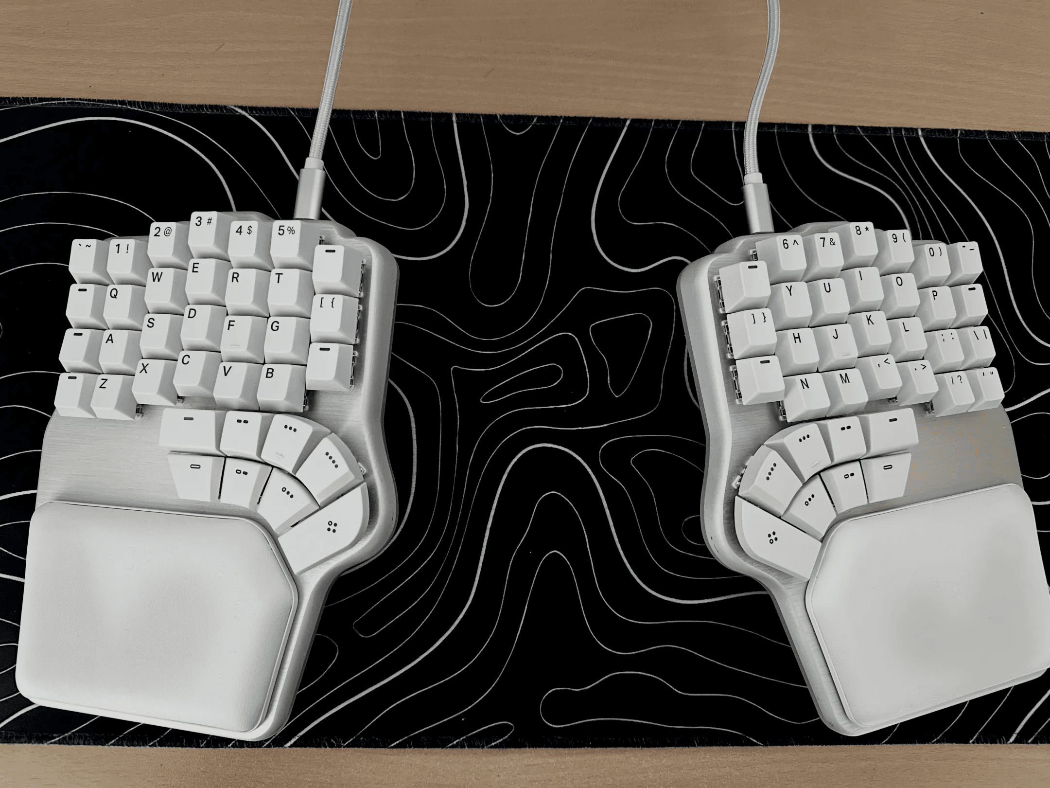 A Dygma Defy split keyboard with white keycaps on a black desk mat with white contour lines, seen from above. Each half has a curved thumb cluster and a padded wrist rest.