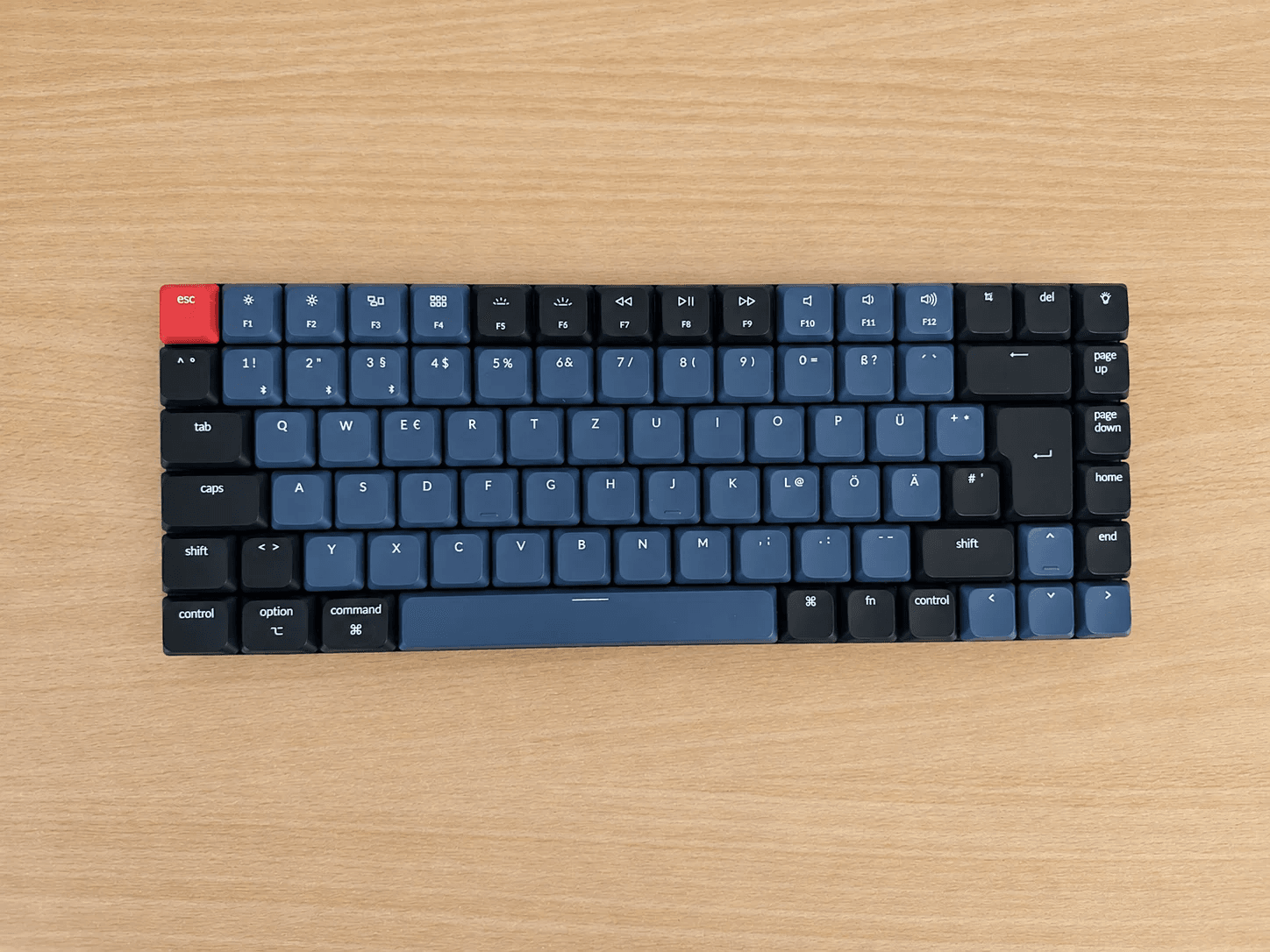A Keychron K3 Pro on a wooden desk, seen from above. It has a German layout with blue and black keycaps, a red Escape key and the legends Ö, Ä, Ü and ß.