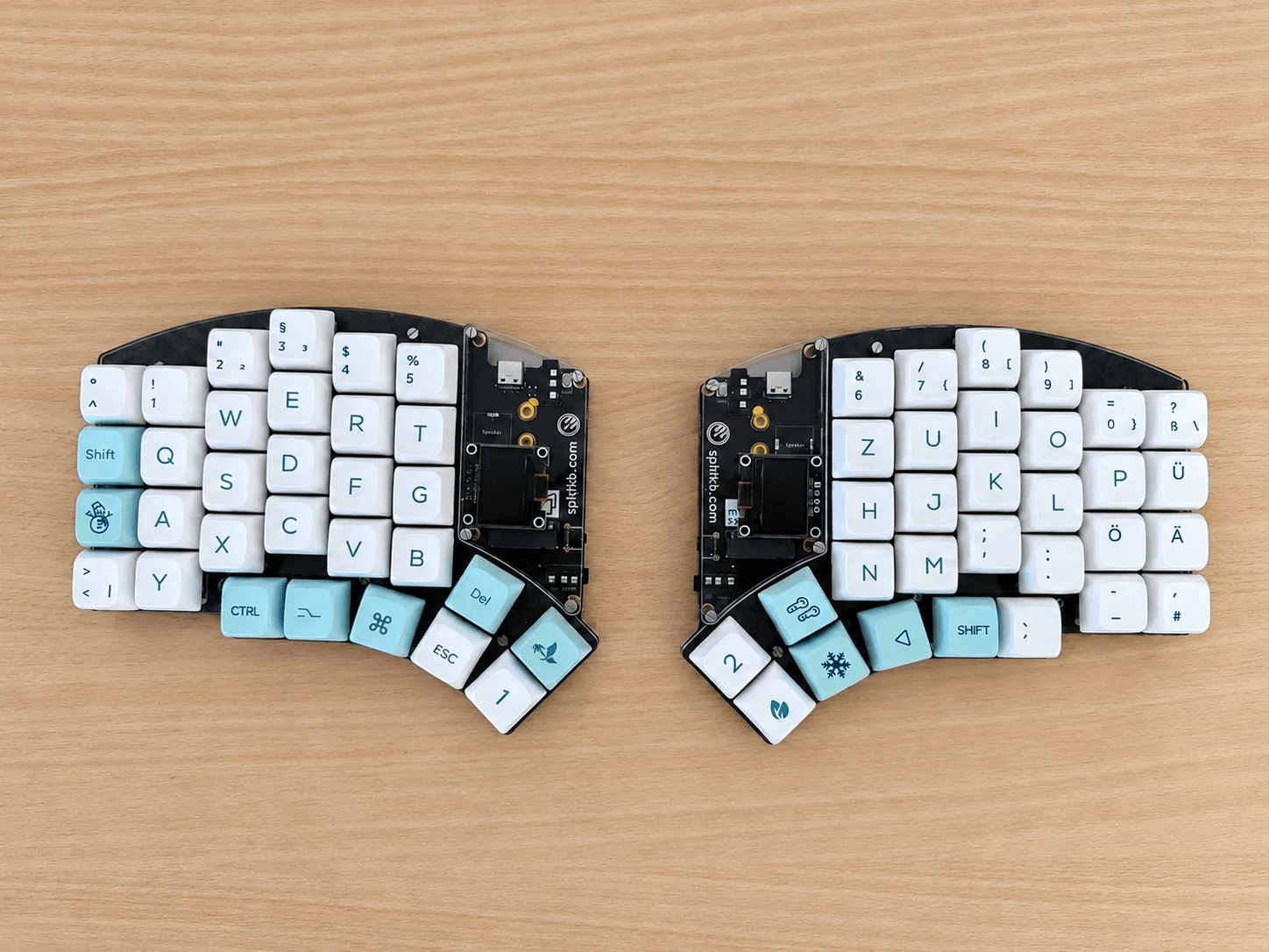 A splitkb Elora split keyboard seen from above on a wooden desk. Each half has a small screen and a thumb cluster, with white and light blue keycaps with German legends.